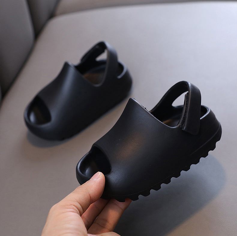 Black children's flip flop sandals with open toes and heel straps, shown on a light gray cushion with one sandal being held by a hand.