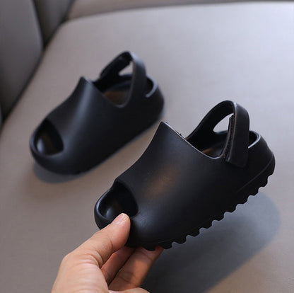 Black children's flip flop sandals with open toes and heel straps, shown on a light gray cushion with one sandal being held by a hand.