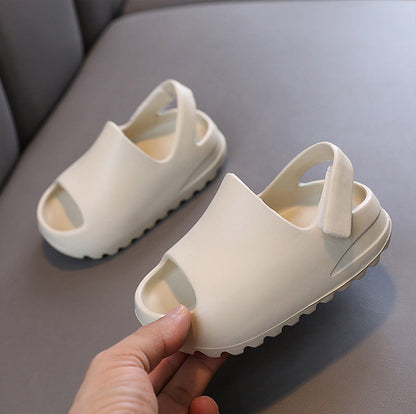 A pair of off-white children's flip-flop sandals with open toes and heel straps, shown on a gray surface with a hand holding one sandal for scale.